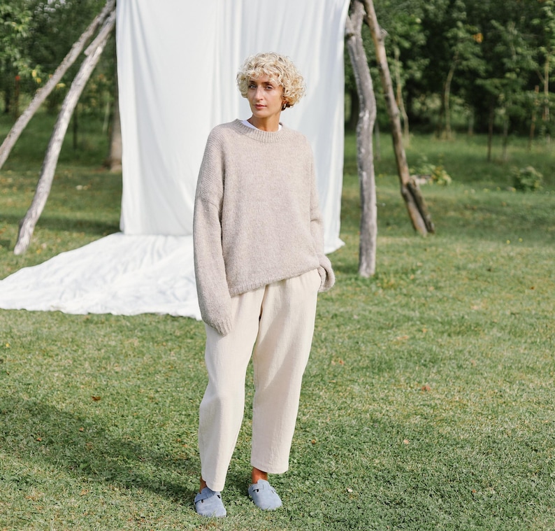 Puede incluir: Una mujer con un su&eacute;ter marr&oacute;n claro y pantalones blancos. Est&aacute; de pie en un &aacute;rea cubierta de hierba con una s&aacute;bana blanca detr&aacute;s de ella.