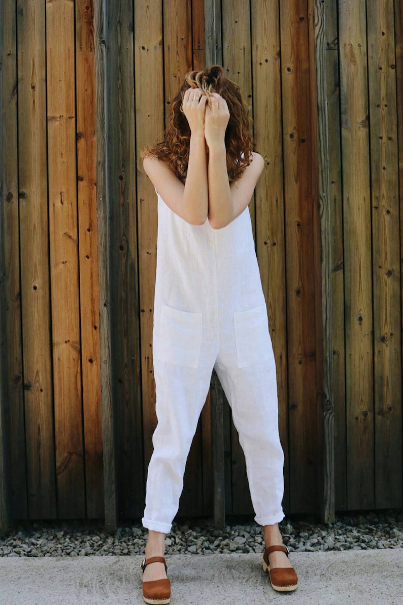 May include: A white sleeveless jumpsuit featuring a round neckline, two large square patch pockets on the front, and tapered pant legs with cuffed hems, paired with brown leather clogs that have a buckle strap and a wooden sole.