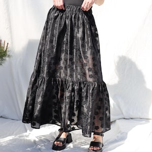 May include: A long, black skirt with a floral pattern and a ruffled hem. The skirt has a high waist and a flowing silhouette. The fabric appears to be slightly sheer, and the skirt is paired with black platform sandals.