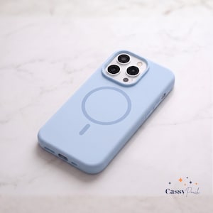 May include: A light blue silicone phone case with a circular MagSafe magnetic charging ring and a vertical alignment line on the back. The case features a square camera cutout with three circular lens openings and the text Cassy Pouch in the corner.