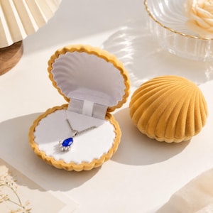 Op de afbeelding: Een mosterdgeel fluwelen sieradendoosje in de vorm van een schelp met een open deksel, met daarin een zilveren ketting met een druppelvormige blauwe edelsteen hanger omringd door kleine heldere steentjes op een witte binnenvoering.
