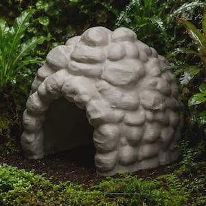 May include: A light gray, dome-shaped animal hideout featuring a textured, stacked stone exterior design. The structure has a single, arched opening at the front, providing a hollow interior space for small pets to rest or seek shelter.