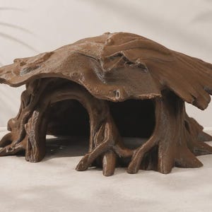 May include: A brown, hollow, tree-stump-shaped shelter with a wide, overhanging roof and a large, arched entrance. The exterior surface features textured, root-like details and a weathered, organic appearance with a matte finish.