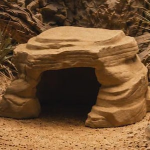 May include: A tan, rock-shaped reptile hide with a textured, layered exterior and a wide, dark arched opening. The structure features a flat top surface and mimics the appearance of natural sandstone, providing a sheltered space for small animals.