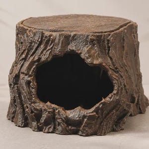 Puede incluir: Una escultura de tocón de árbol hueco de color marrón con un exterior texturizado similar a la corteza y una gran abertura oscura e irregular en la parte frontal. La superficie superior es plana y circular, imitando un tronco cortado.