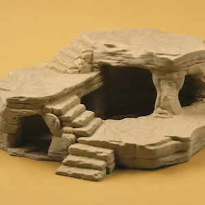 May include: A beige, rock-like structure featuring a multi-level design with carved stairs leading to an upper platform. The object includes a hollow cave-like opening underneath the elevated section, with textured surfaces mimicking natural stone formations.