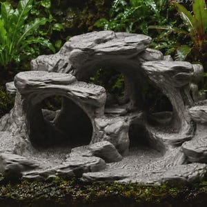 Puede incluir: Una estructura de formación rocosa gris de varios niveles que presenta varias aberturas de cuevas huecas y plataformas escalonadas. La superficie de la estructura muestra una apariencia texturizada y en capas con detalles tallados que imitan la piedra.