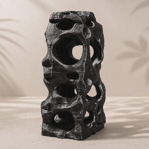 May include: A tall, vertical, black sculpture with a rough, textured surface and multiple irregular, hollow circular openings throughout its structure. The object features a weathered, stone-like appearance with subtle grey highlights on the edges.