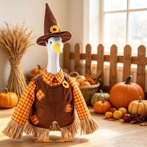 May include: A white goose figurine dressed as a scarecrow, wearing a brown pointed hat with a sunflower, a plaid orange and yellow shirt, and brown overalls with plaid patches. The base features straw-like fringe and orange webbed feet.