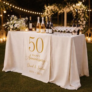 May include: A cream-colored table runner featuring gold text that reads 50 HAPPY Anniversary David & Sophia 1975 - 2025. The runner is draped over a table set with wine bottles, clear glasses, and a floral arrangement in an outdoor evening setting.
