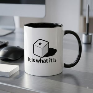 May include: A white ceramic mug with a black interior and handle. The front features a black graphic of a single die showing one dot, accompanied by the text "It is what it is" printed in a bold, black, sans-serif font below the die illustration.