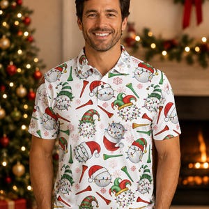 May include: A white short-sleeve polo shirt featuring an all-over print of golf balls wearing various holiday hats, including Santa hats and elf caps, wrapped in colorful string lights, interspersed with red and green golf tees and snowflakes.