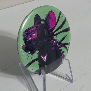 May include: A circular pin-back button featuring a black cybernetic mouse illustration on a light green background. The mouse wears a digital headset with a pink screen, has glowing pink accents, and is connected to multiple black cables.