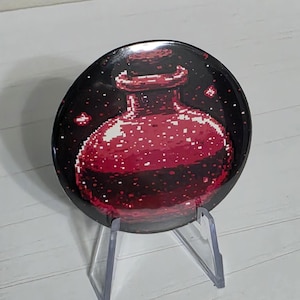 May include: A circular pinback button featuring a pixel art illustration of a round glass bottle filled with a dark red liquid. The bottle is set against a black background with small, scattered white pixelated sparkles or light reflections.