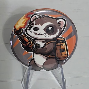 May include: A circular pin-back button featuring a cartoon ferret with dark brown fur and a white chest. The ferret holds a flamethrower with a bright orange flame and wears a tan backpack. The background is a circular design with orange and gray stripes.