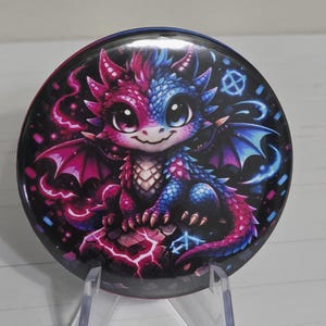 May include: A circular pinback button featuring a vibrant illustration of a small dragon split down the middle with pink and blue scales. The dragon has large eyes, wings, and sits on a rocky surface against a dark background with glowing accents.