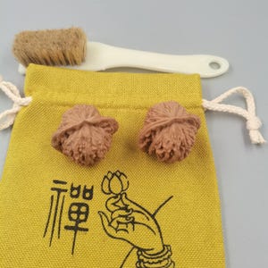 May include: A mustard yellow drawstring pouch featuring a black graphic of a hand holding a lotus flower and the Chinese character for Zen. Two brown, textured walnut shells rest on the pouch, accompanied by a white cleaning brush with tan bristles.