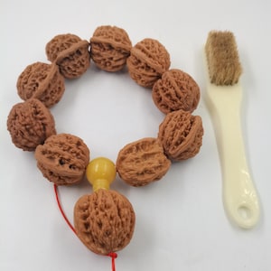 May include: A circular string of ten brown, textured walnut shells connected by a red cord, featuring a single smooth yellow bead near the bottom. Beside the walnut string lies a small cleaning brush with a cream-colored handle and brown bristles.