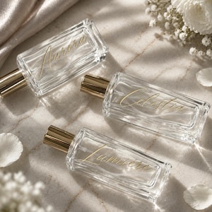 May include: Three rectangular clear glass perfume bottles with gold-toned caps. Each bottle features elegant gold cursive lettering on the front, displaying the names Aurora, Celestia, and Lumière respectively, arranged in a diagonal composition.