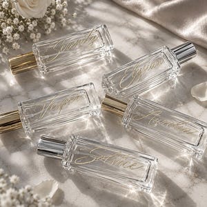 Puede incluir: Cinco frascos de perfume rectangulares de vidrio transparente con tapas metálicas. Cada frasco presenta un texto dorado en cursiva que dice Serenity, Elysian, Vesper, Aurelia y Solstice. Los frascos están dispuestos en un grupo.