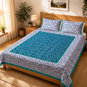 Könnte beinhalten: Eine Tagesdecke mit einem zentralen rechteckigen Feld mit blau-türkisfarbenem Mandala-Muster, umgeben von einem weißen Rand mit blauem Rankenmuster. Zwei passende rechteckige Kopfkissen liegen am Kopfende des Bettes.