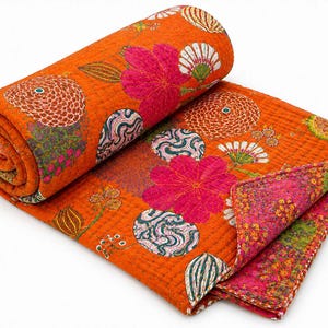 Könnte beinhalten: Eine gerollte Steppdecke mit leuchtend orangefarbenem Grund und einem wiederkehrenden Muster aus großen rosa Blumen, kreisförmigen Motiven mit geschwungenen Linien und kleinen botanischen Elementen. Die Unterseite zeigt einen dichten Blumendruck.