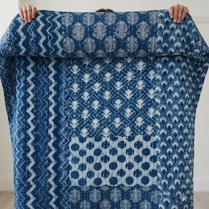 Può includere: Una trapunta rettangolare con un design patchwork composto da vari motivi blu e bianchi, tra cui zig-zag, disegni paisley, rombi floreali e archi smerlati, rifinita con cuciture bianche a vista su tutta la superficie.