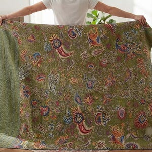 Op de afbeelding: Een rechthoekige quilt met een olijfgroene basiskleur en een dicht, veelkleurig bloemen- en pauwenpatroon. De stof vertoont overal ingewikkelde stiksels, met een effen olijfgroene rand aan de linkerkant van het textiel.