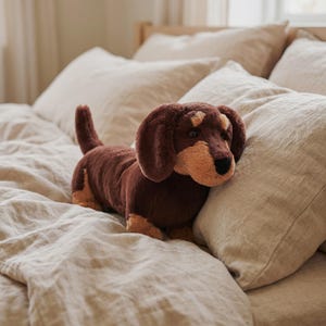 May include: A plush dachshund toy featuring dark brown fur on the body and ears, with lighter tan accents on the muzzle, above the eyes, and on the paws. The stuffed animal has a long body, a short tail, and a black nose with dark eyes.