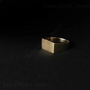May include: A gold-toned signet ring featuring a flat, rectangular top surface with a matte finish. The band is smooth and tapers toward the back. The text Luxe Diam Jewel is visible in the background, partially overlapping the ring's position.