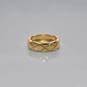 May include: A polished gold band ring featuring a repeating quilted diamond pattern across the outer surface. The ring has a smooth, reflective finish with scalloped edges along the top and bottom, creating a textured, geometric design.