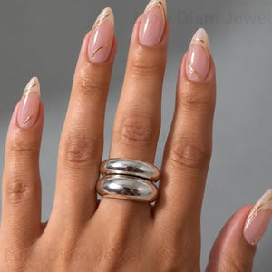 May include: Two stacked silver-toned rings with a smooth, polished, rounded surface worn on a finger. The fingernails are shaped into a long almond style, featuring a light pink base colour accented with delicate, thin, wavy gold-coloured lines.