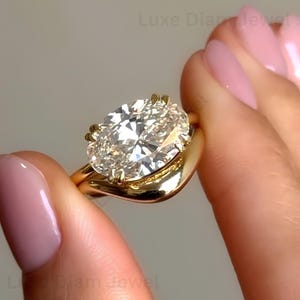 Peut inclure: Une bague en or avec une grande pierre précieuse transparente taille ovale à facettes, fixée par quatre griffes. L'anneau présente une finition lisse et polie avec une forme légèrement ondulée. Le texte Luxe DiamJewel est visible.