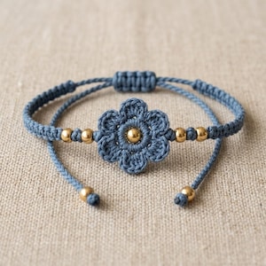 May include: A blue macrame bracelet featuring a central crocheted flower motif with a gold-toned bead at its center. The cord is woven with additional gold-toned beads on either side of the flower and at the ends of the adjustable pull strings.