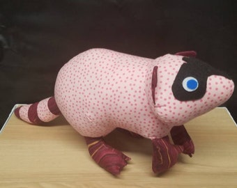 Könnte beinhalten: Ein rosafarbenes und rotes Plüsch-Waschbär-Spielzeug mit schwarzen Augen und einer schwarzen Nase. Das Spielzeug sitzt auf einer Holzoberfläche.