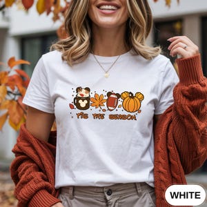 May include: A white short-sleeved t-shirt featuring a graphic print of a brown mug, an orange maple leaf, a brown football, and an orange pumpkin, all with mouse ear silhouettes. The text 'TIS THE SEASON' is printed in brown below the icons.
