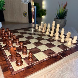 Könnte beinhalten: Ein hölzernes Schachspiel mit einem klappbaren Brett in einem Schachbrettmuster aus dunkelbraunem Maserholz und hellem Creme. Das Set enthält einen vollständigen Satz dunkelbrauner und cremefarbener Holzschachfiguren in Startposition.