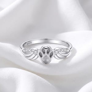 Könnte beinhalten: Ein silberfarbener Ring mit einem zentralen Pfotenabdruck-Motiv, das von zwei Flügeln flankiert wird. Die Flügel sind mit kleinen, klaren, funkelnden Steinen besetzt. Die Innenseite des Rings ist deutlich mit S925 geprägt, was die Zusammensetzung angibt.