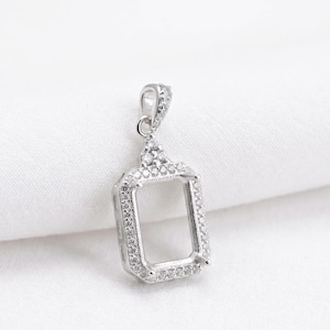 May include: A silver-toned rectangular pendant setting featuring a hollow center framed by small, clear, sparkling stones. The pendant includes a decorative bail at the top, also accented with small stones, designed to hold a gemstone or insert.