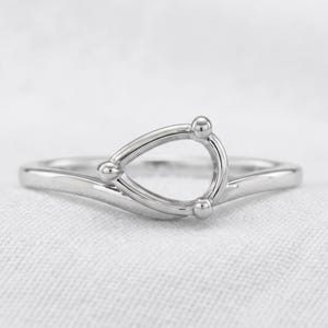 Könnte beinhalten: Ein polierter silberfarbener Ring mit einer tropfenförmigen, offenen Fassung. Der tropfenförmige Rahmen ist mit drei kleinen, kugelförmigen Metallperlen an den Eckpunkten verziert, was dem Ring ein klares und strukturelles Design verleiht.