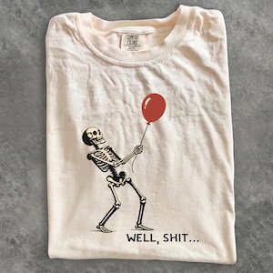 Peut inclure: Un t-shirt couleur crème avec un motif noir représentant un squelette tenant un ballon rouge. Sous le squelette, le texte indique "WELL, SHIT...". Le t-shirt présente une encolure ronde et une étiquette de marque Comfort Colors à l'intérieur.