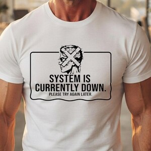 May include: A white short-sleeved t-shirt featuring a black graphic of a human head with an X over the face. Below the graphic, the text reads: SYSTEM IS CURRENTLY DOWN. PLEASE TRY AGAIN LATER. The design is enclosed within a rectangular border.