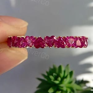 May include: A gold band ring featuring a continuous row of vibrant, deep pink heart-shaped gemstones. The stones are set side-by-side in a prong setting, creating a uniform line of faceted hearts that catch the light across the entire circumference.