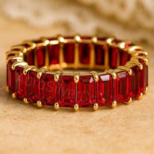 Op de afbeelding: Een gouden eeuwigheidsring met een doorlopende rij levendige rode edelstenen in smaragdgeslepen vorm, gezet in een gepolijst gouden frame. De ring toont een herhalend patroon van rechthoekige stenen die door kleine gouden klauwtjes worden vastgehouden.