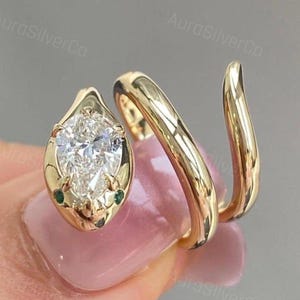 Könnte beinhalten: Ein goldfarbener Wickelring mit einem birnenförmigen, klaren Edelstein an einem Ende und einer glatten, sich verjüngenden Ringschiene, die sich um den Finger legt. Die Fassung enthält zwei kleine grüne Akzentsteine an der Basis des Hauptsteins.