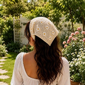 May include: A triangular crochet headscarf in a light cream color featuring a repeating pattern of white daisy motifs with yellow centers. The accessory is crafted with an open-knit texture and is tied at the back of the head to cover the hair.