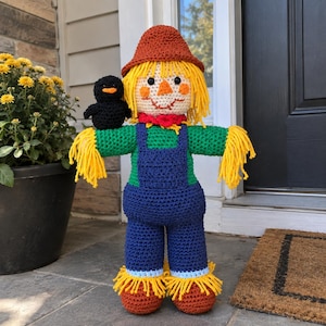May include: A crocheted scarecrow doll featuring a rust-colored hat, yellow yarn hair, and a black bird perched on its shoulder. The doll wears a green long-sleeve shirt, blue overalls, and brown shoes with yellow fringe accents at the wrists and ankles.