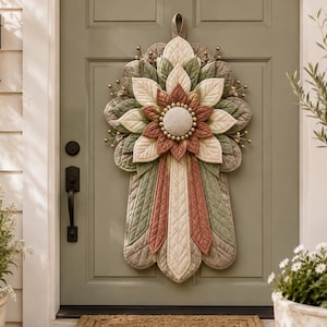 Peut inclure: Une décoration de porte en tissu matelassé présentant un motif floral superposé dans des tons vert sauge, crème et terre cuite. L'élément circulaire central est entouré de segments en forme de pétales avec des détails cousus et des accents dorés.