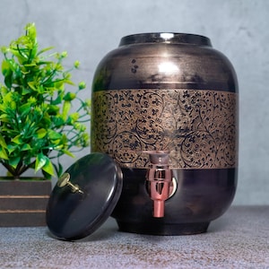 Puede incluir: Un dispensador de bebidas de metal oscuro con acabado en bronce cepillado y una banda central ancha con un intrincado patrón floral. El recipiente cilíndrico incluye una tapa a juego con un pequeño pomo y un grifo metálico frontal.