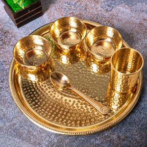 May include: A gold-colored metal dinnerware set consisting of a large round tray, three small bowls, a tall tumbler, and a spoon. Each piece features a hammered texture and a polished finish. The tray has a beaded edge, and the spoon handle has a decorative floral pattern at the end.